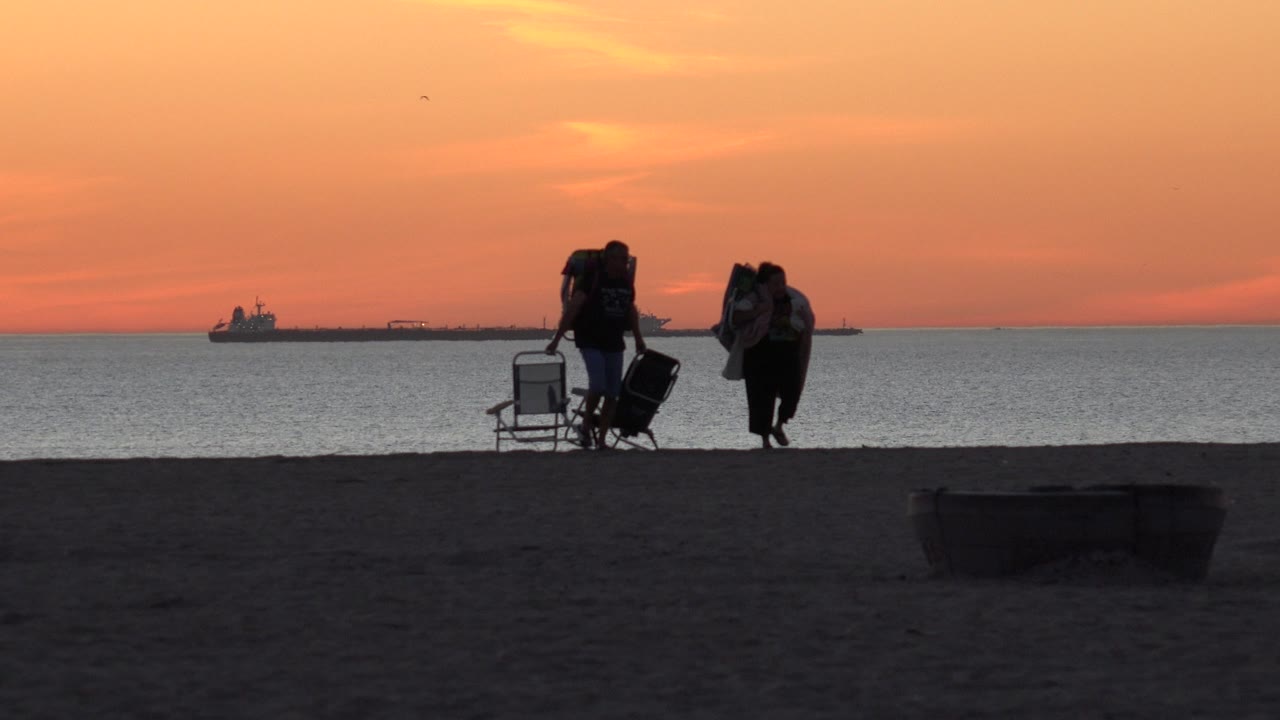 Two silhouetted people with beach gear pass in front of the ocean.