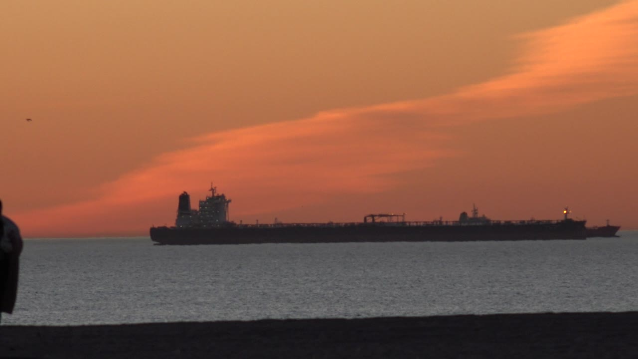A cargo ship is silhouetted on the ocean at Huntington Beach, California, while a cropped person stands near the left foreground edge under warm orange cloud color.