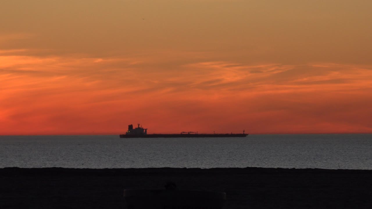 Opening frame of a cargo ship silhouetted off Huntington Beach at orange sunset, with dark shoreline in the foreground and no visible people.