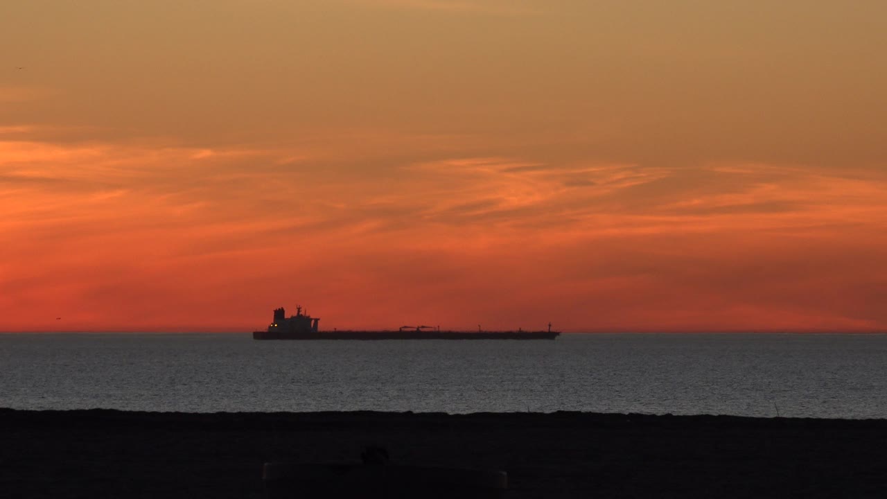 A tanker silhouette sits near the Huntington Beach ocean horizon beneath orange sunset clouds, with a dark shoreline in the foreground and no visible people.