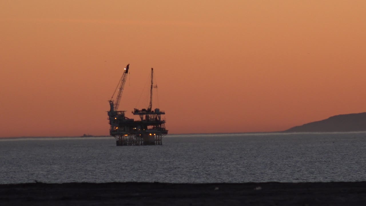 At the start of the Huntington Beach clip, an offshore platform with cranes sits left of center across dark water under an orange dusk sky, with no visible people.