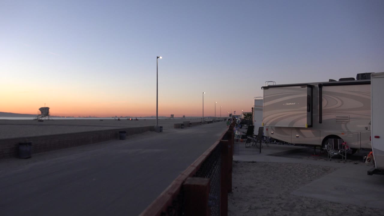 Huntington Beach dawn scene showing a broad paved path, a sandy edge, RVs parked on the right, a line of tall lamps, and no visible people.