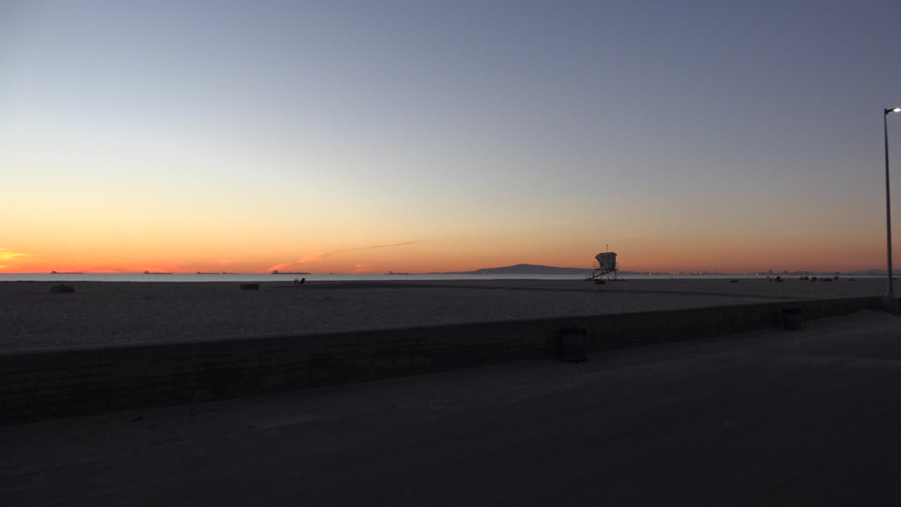 Huntington Beach dusk view with a dark foreground wall, open sand, a tall light pole near the right side, a distant lifeguard tower shape, orange horizon light, and no visible people.