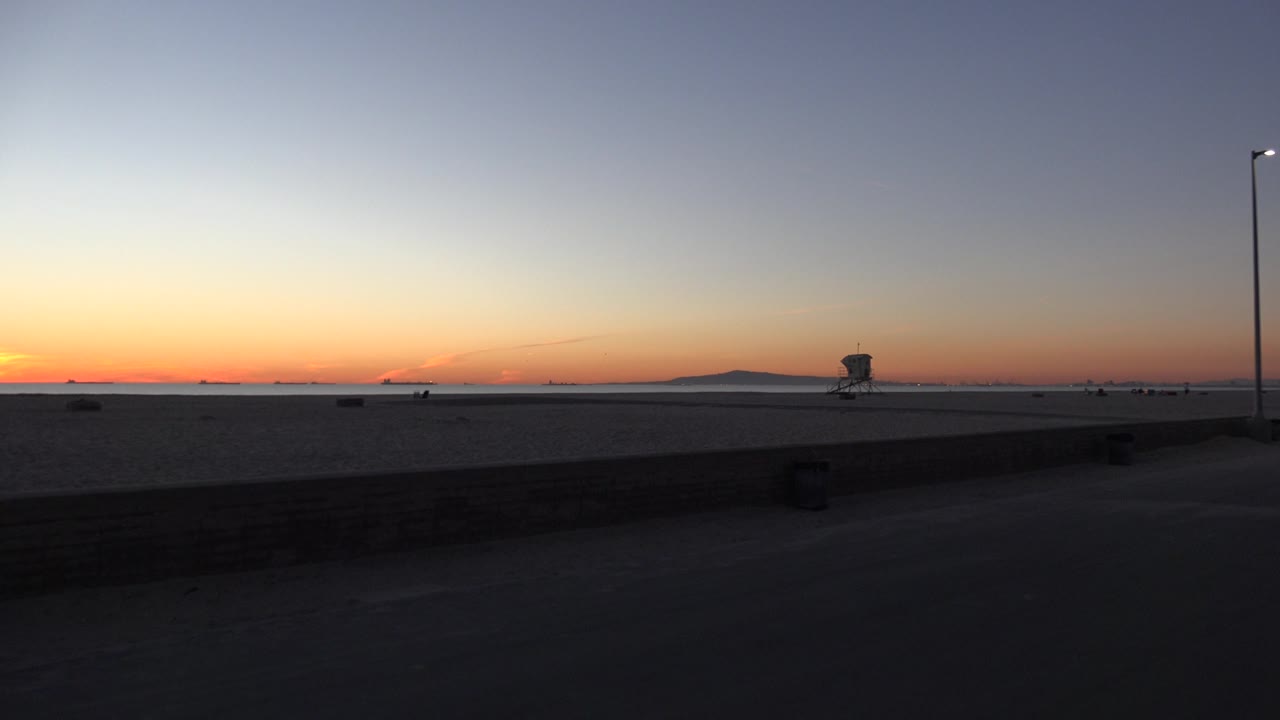 Orange dusk sky over beach, ocean, light pole, and distant tower.