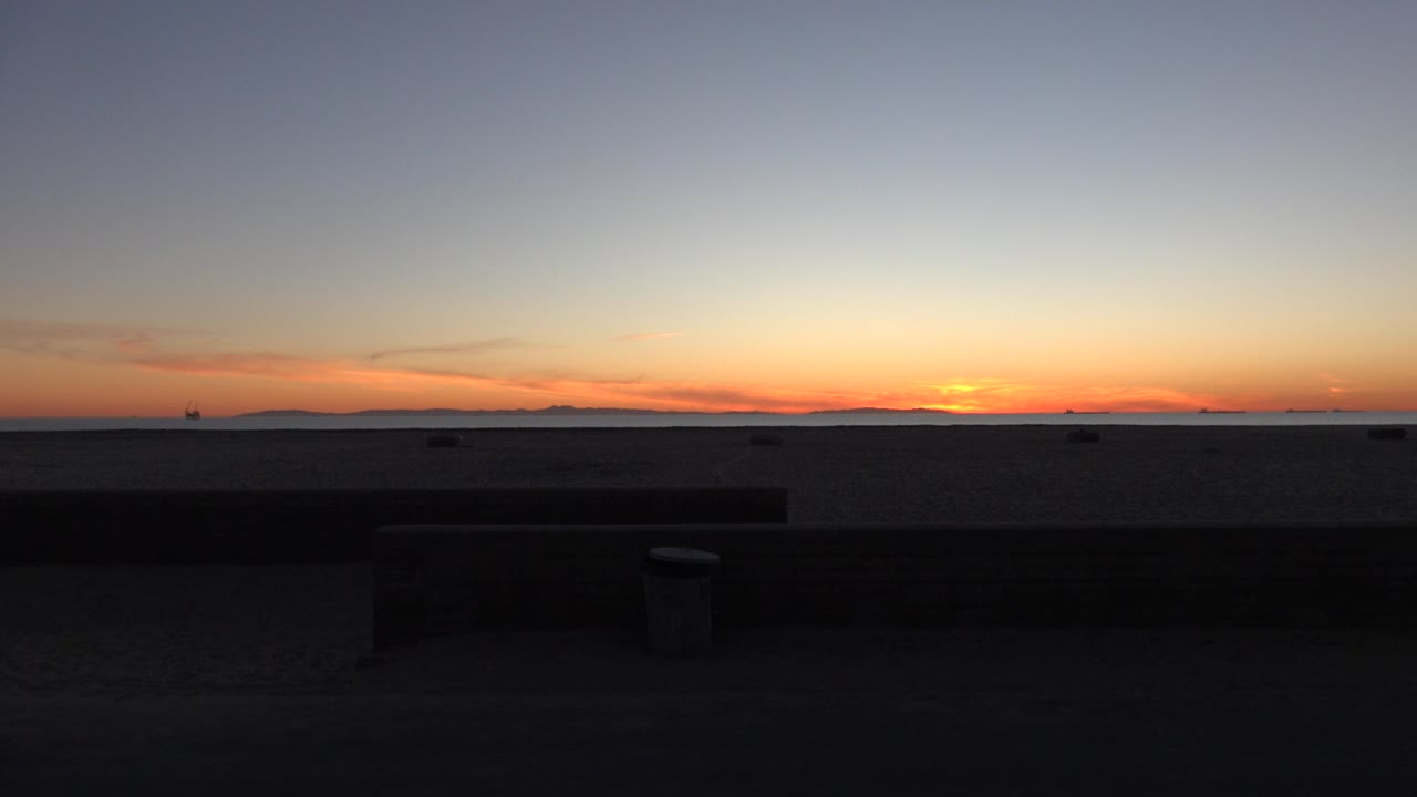 Opening frame of a Huntington Beach ocean sunset with a dark low wall or bench in front, distant land on the horizon, orange light near the water, and no visible people.