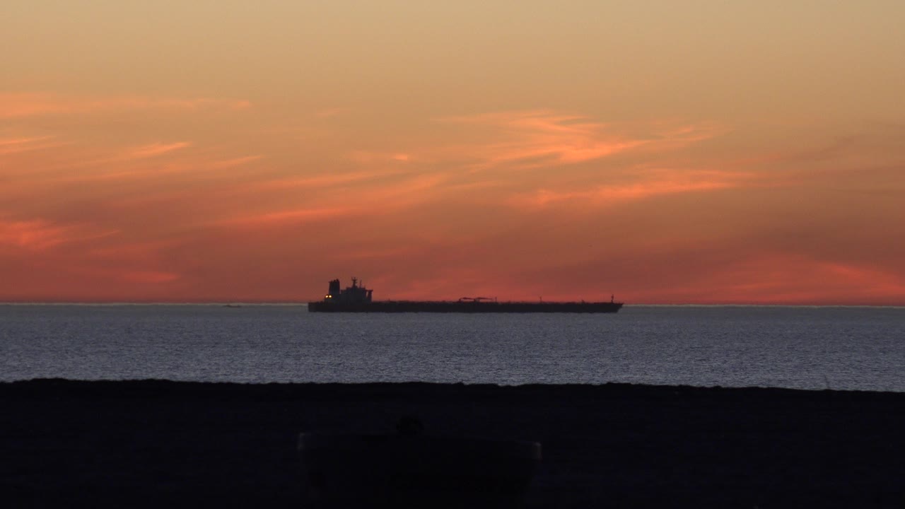 At 0:00, a distant cargo ship sits left of center on the Huntington Beach ocean horizon beneath orange sunset clouds, with dark foreground and no visible people.
