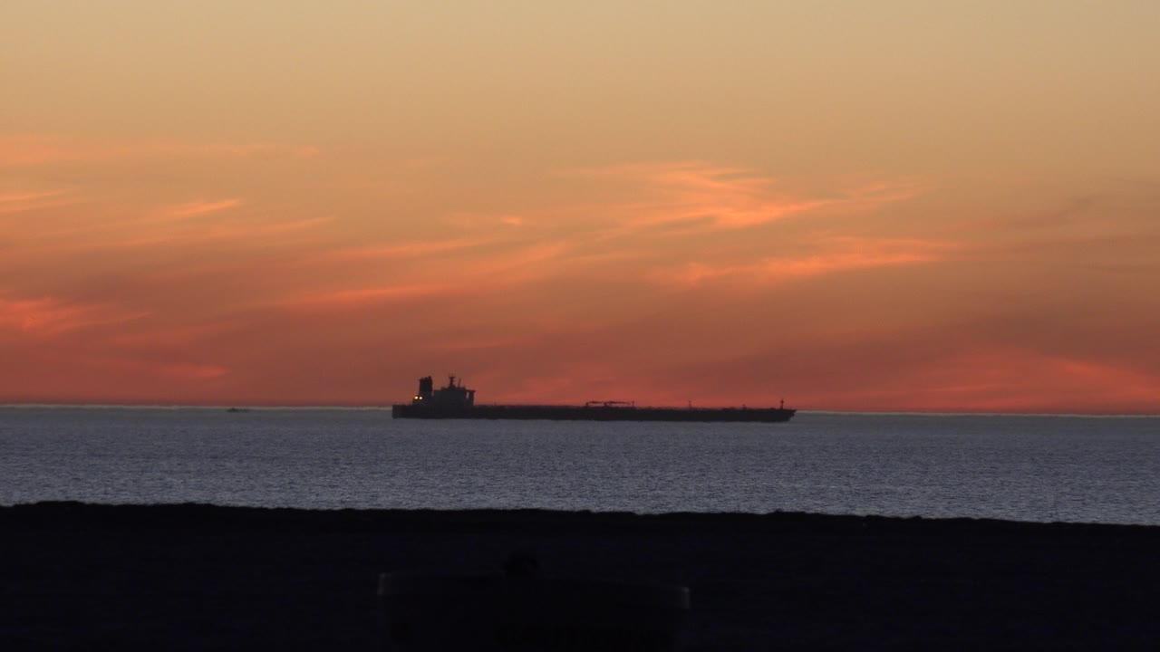 At 0:01 in Huntington Beach, California, the tanker remains small and dark on the horizon while red-orange cloud bands deepen above the water, with no visible people.