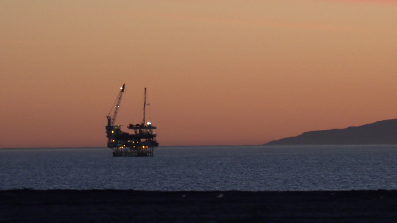 Huntington Beach ocean view with a dark offshore structure and crane arms left of center, orange sunset sky, distant land at right, and no visible people.