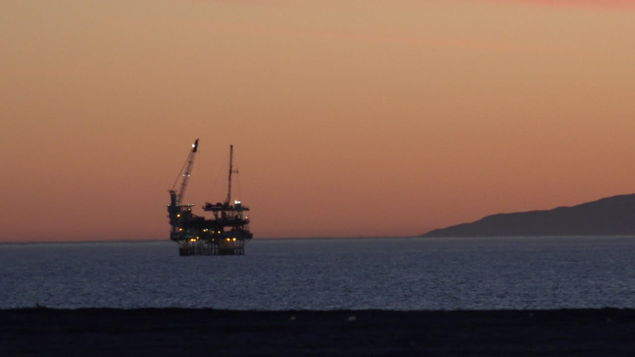 At 0:00, a distant offshore structure with cranes and small lights sits off Huntington Beach under an orange sky, with dark beach foreground and no visible people.
