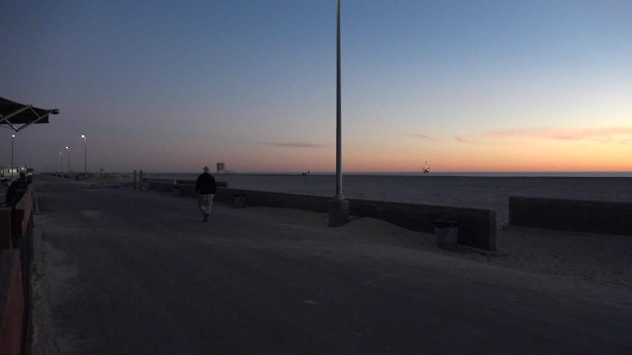 Dusk beach promenade with a person walking near a wall and ocean horizon.