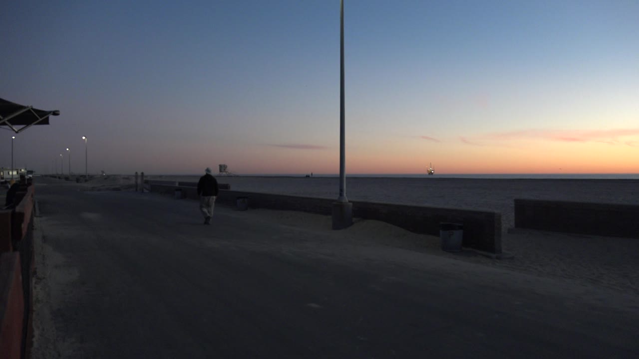 At 0:01 in Huntington Beach, the walker is slightly farther down the promenade, framed by the low wall, sand, ocean, and warm sunset band on the horizon.