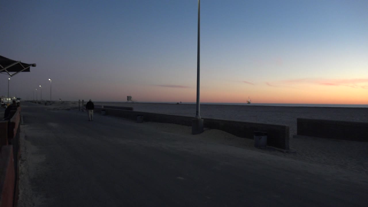 A lone figure continues along the Huntington Beach shoreline as the foreground stays dark, the ocean lies to the right, and a warm sunset band brightens the horizon.