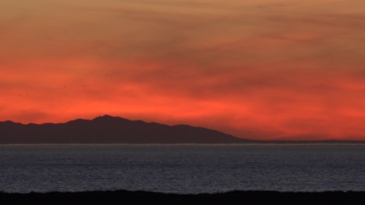 Red sunset sky over ocean with distant dark land silhouette.
