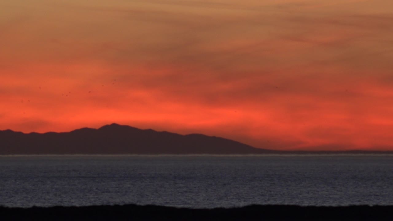At 0:01 in Huntington Beach, the distant dark landform sits lower beneath a broad red cloud band over the ocean, with no visible people.