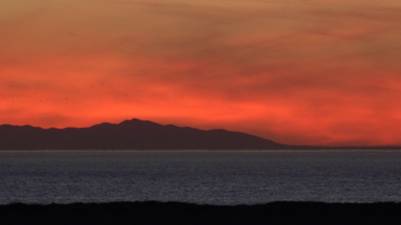 Red sunset sky over ocean with distant dark land silhouette.