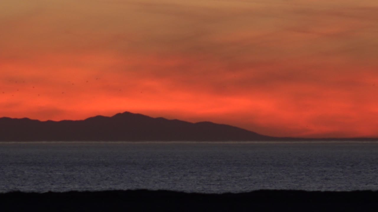 At 0:01 in Huntington Beach, California, the horizon centers a dark coastline shape below a red cloud layer, with ocean texture in the midground and no visible people.