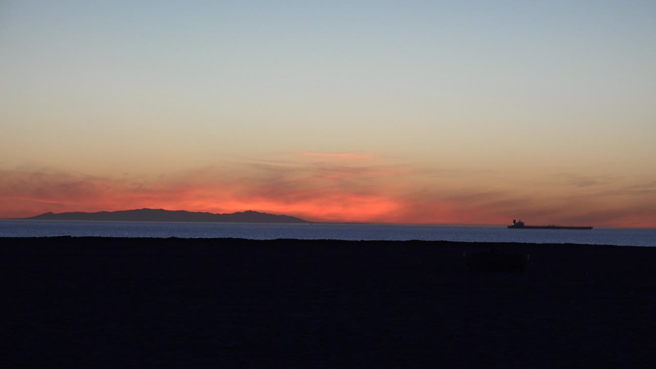Huntington Beach sunset over the ocean with a distant tanker near the right horizon, dark beach foreground, orange cloud glow, and no visible people.