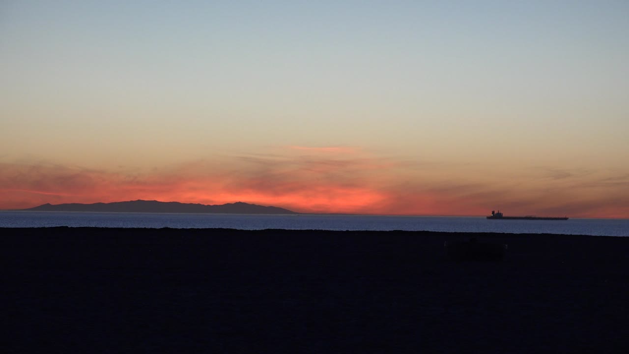 A small bird appears above the low ocean horizon at Huntington Beach while a tanker sits offshore under orange sunset clouds, with no visible people.
