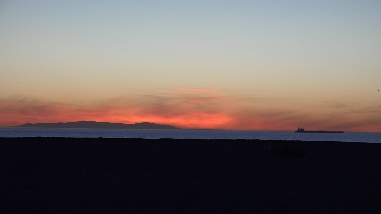 Orange sunset glow over ocean with a distant tanker and dark foreground.