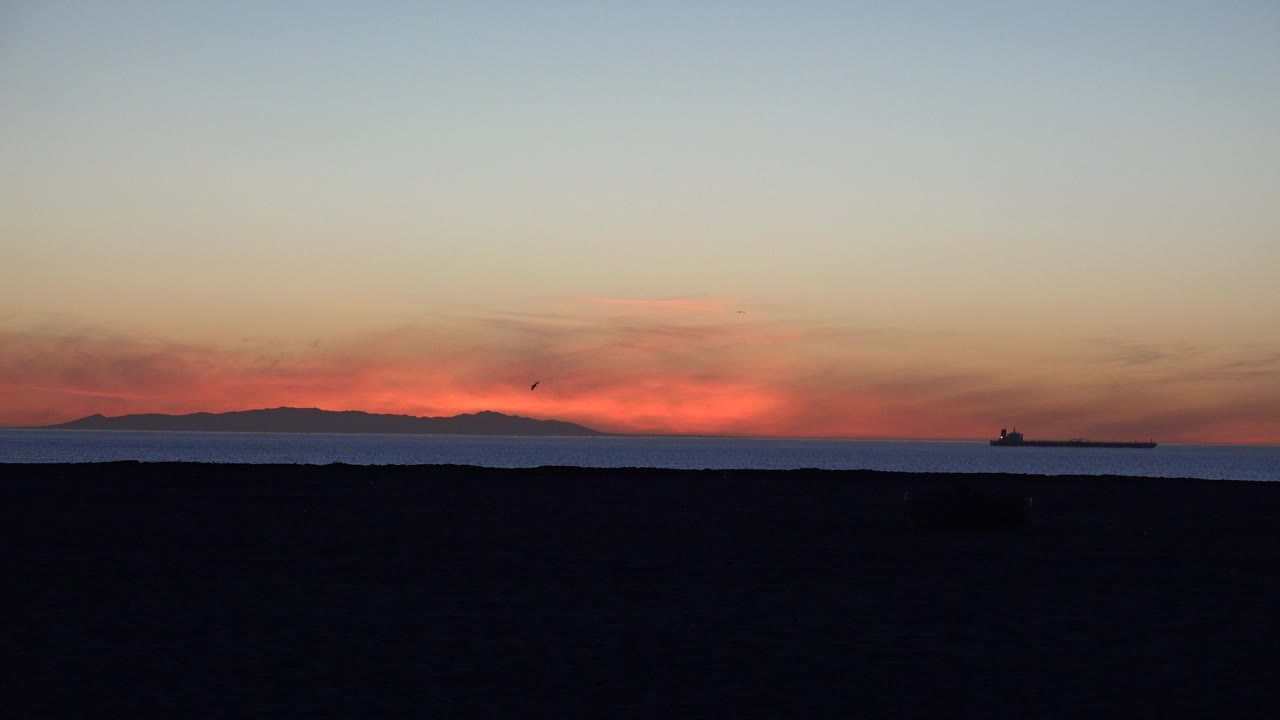 Sunset over ocean with dark beach foreground and distant ship.