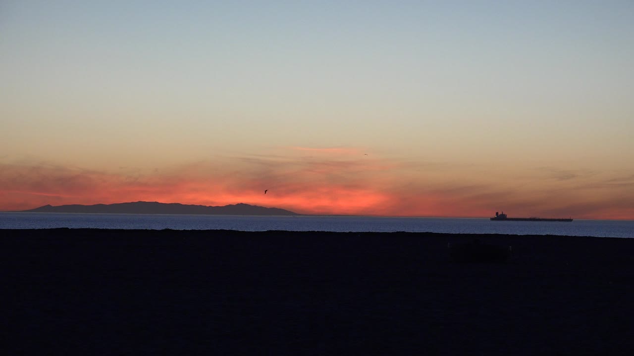 Huntington Beach ocean sunset with the ship sitting near the right side of the horizon, red light under thin clouds, dark sand below, and no visible people.