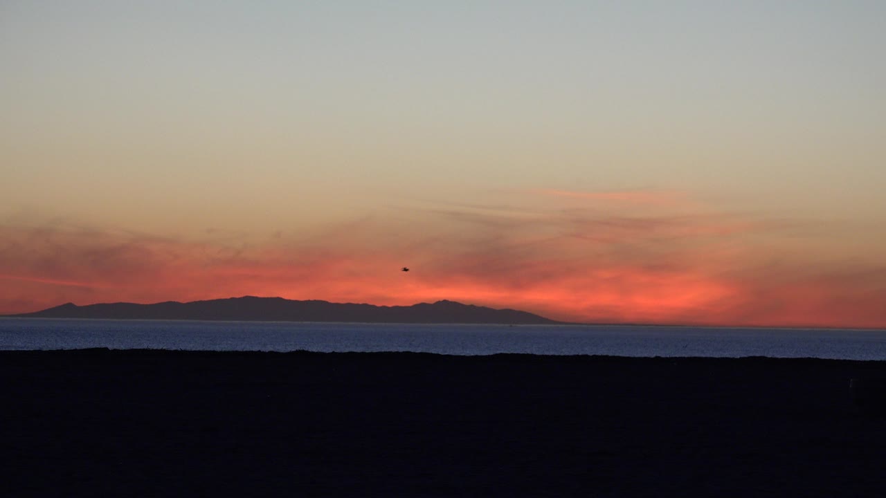 Opening frame of Huntington Beach at dusk with a dark beach foreground, blue ocean band, distant land on the horizon, orange cloud glow, and no visible people.