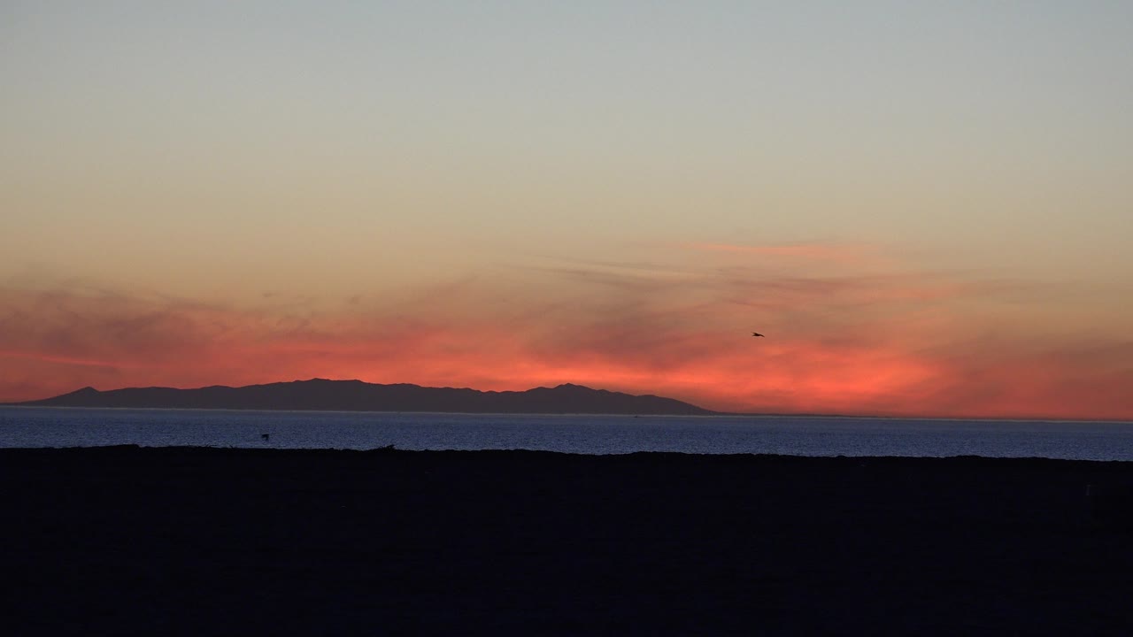Opening frame of Huntington Beach with a dark beach foreground, blue ocean band, distant landform on the horizon, red-orange sky glow, and no visible people.