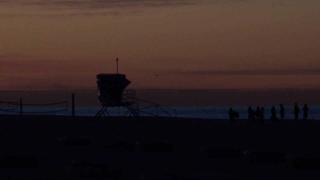 At Huntington Beach, silhouetted people gather on dark sand beside a beach tower, with low waves and a warm sunset sky behind them.