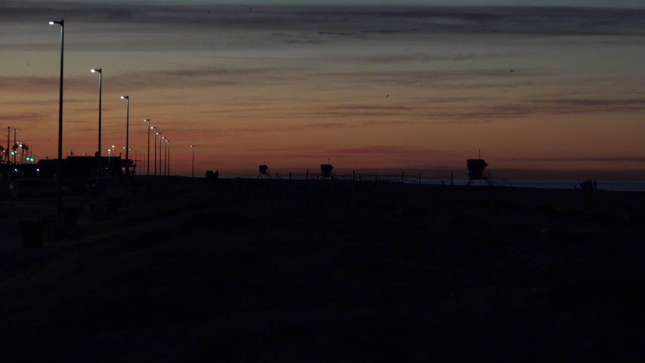 Huntington Beach harbor at dusk with a dark foreground, lit streetlights along the left side, a low ocean horizon, layered sunset color, and no visible people.