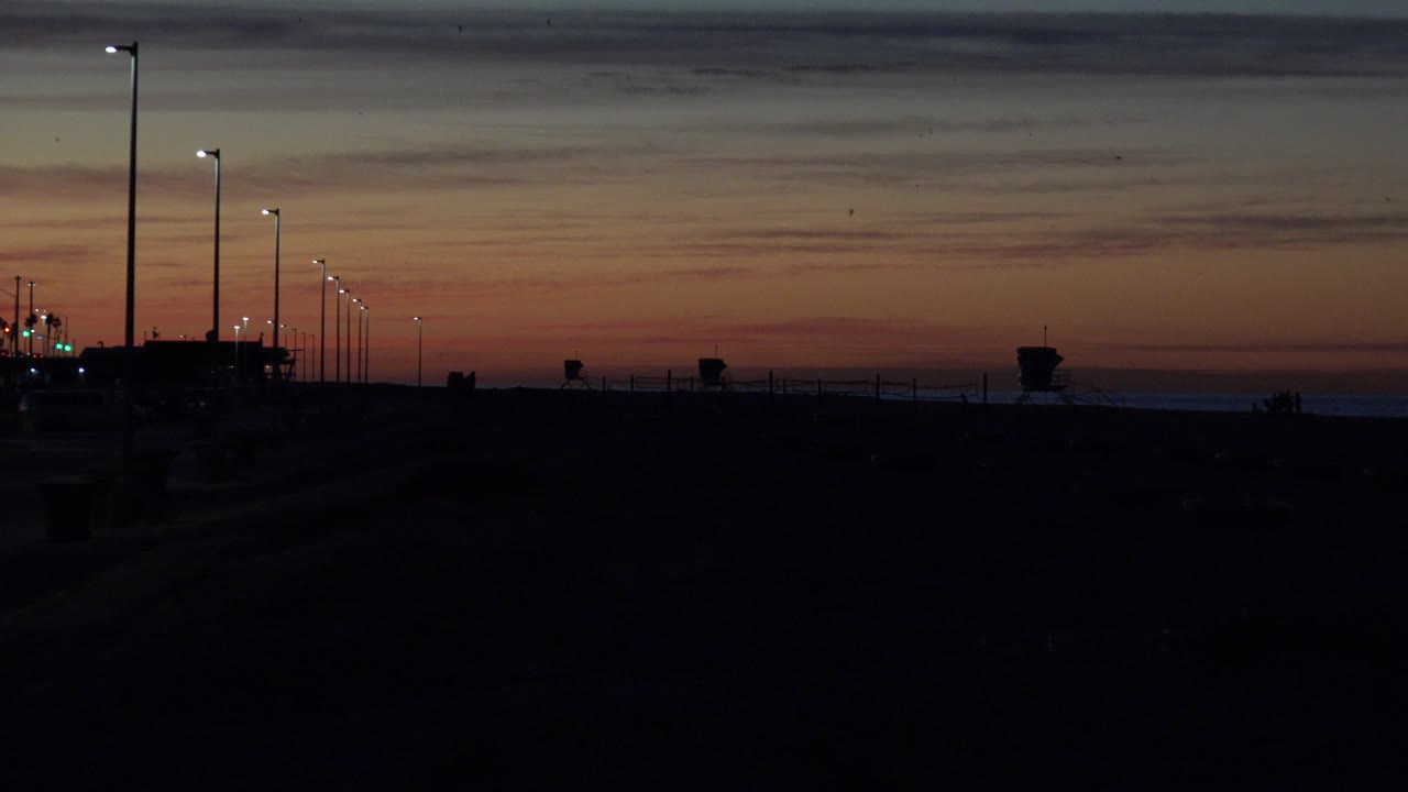 Huntington Beach sunset view with streetlights receding into the distance, dark harbor shapes in the foreground, a narrow ocean line, and no visible people.