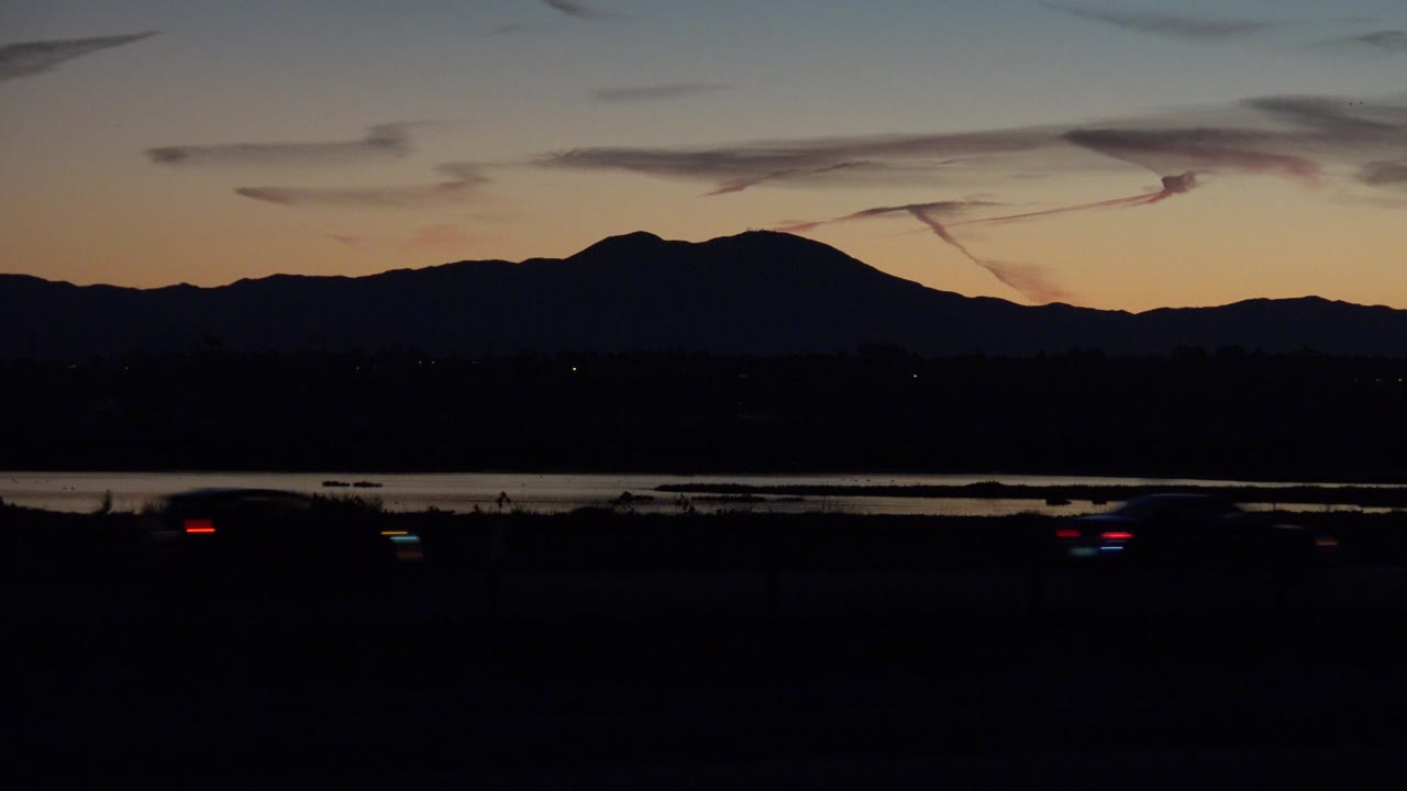 At 0:00, Huntington Beach, California dusk shows a dark roadway foreground, a thin water band, mountain silhouettes, and an orange-blue sky with no visible people.