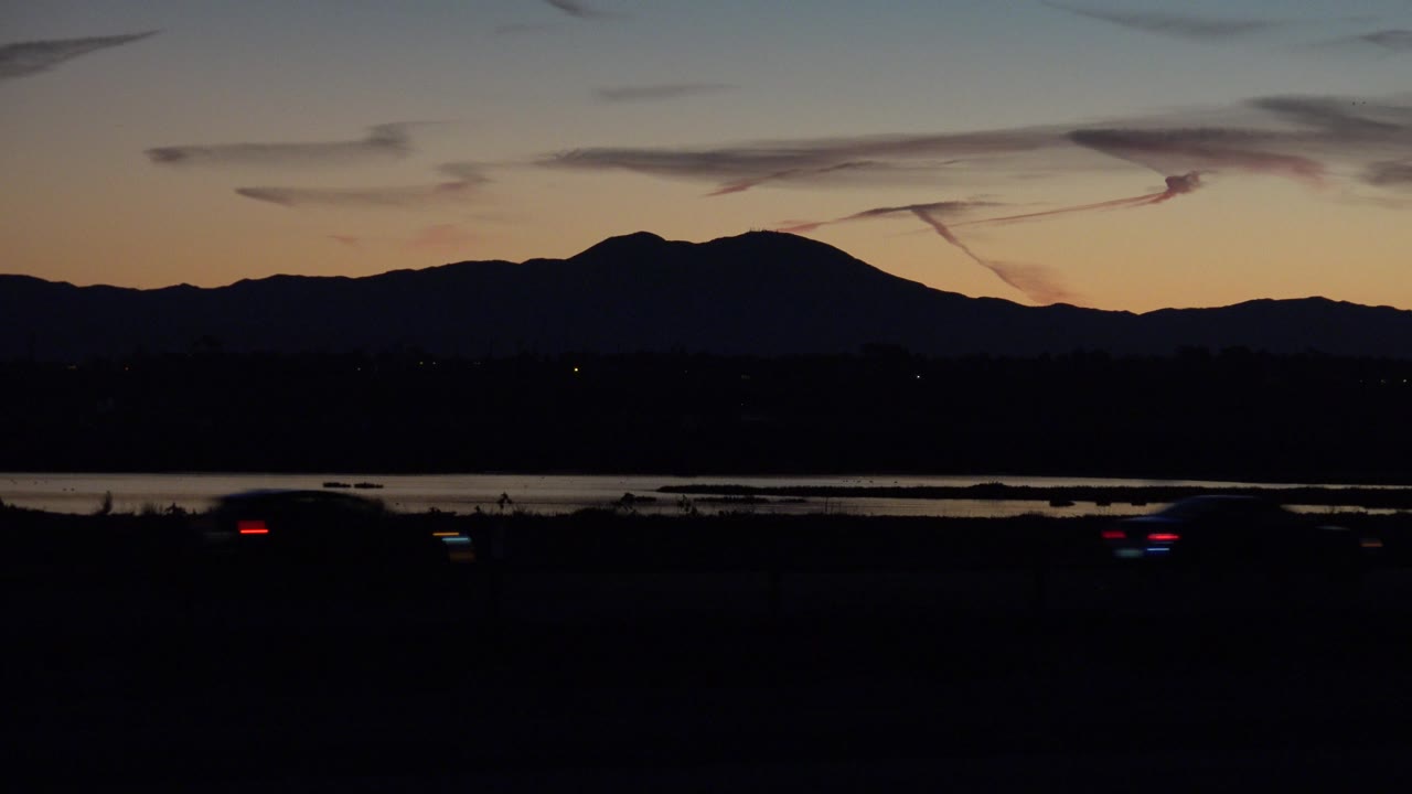 At 0:01, Huntington Beach, California appears as a low dark highway with small red and white vehicle lights, distant water, and a broad mountain ridge under fading sky, with no visible people.