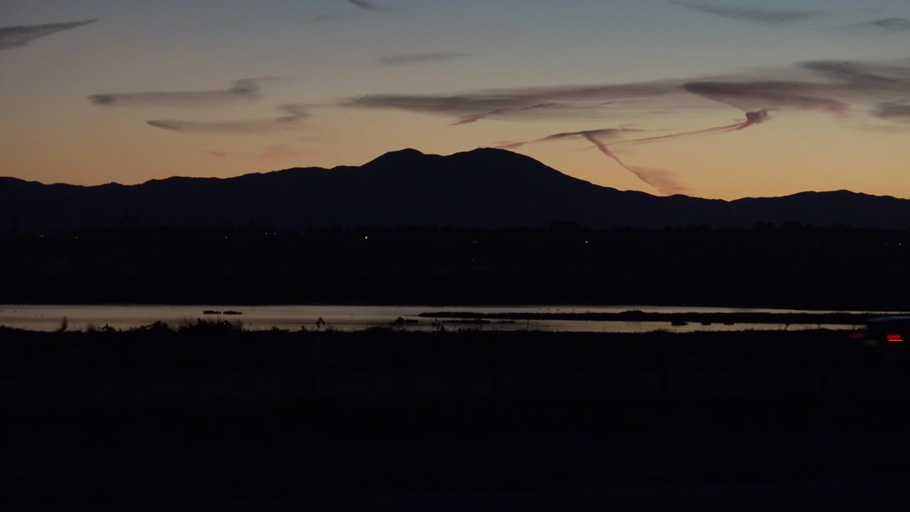 Twilight mountain skyline with a dark road and small vehicle lights.