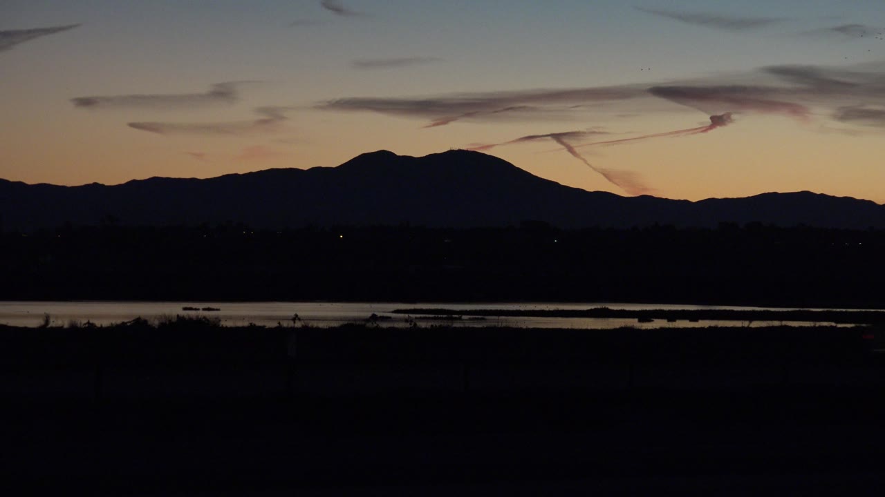 Huntington Beach twilight view with a dark highway foreground, a narrow water band, silhouetted mountains, orange-blue sky, and no visible people.