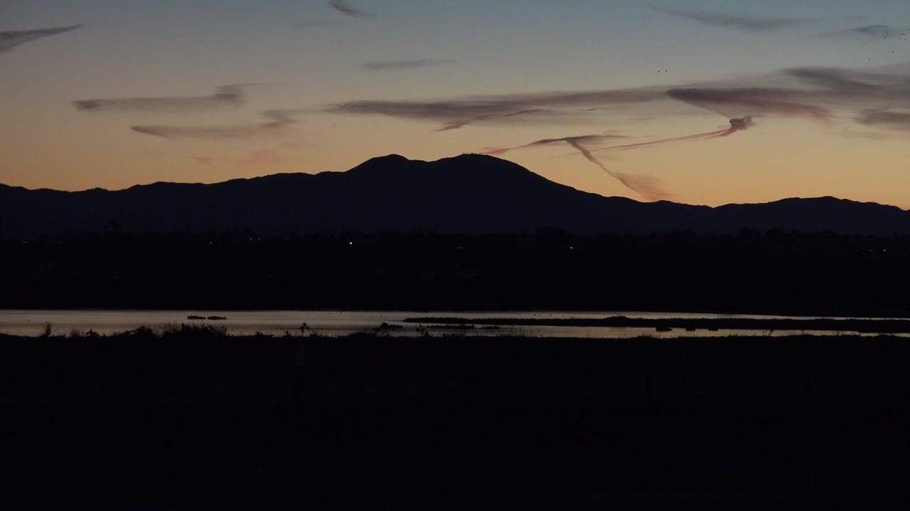 Huntington Beach sunset frame with the mountain ridge centered, water reflecting a thin line of light, a dark road at the bottom, and no visible people.