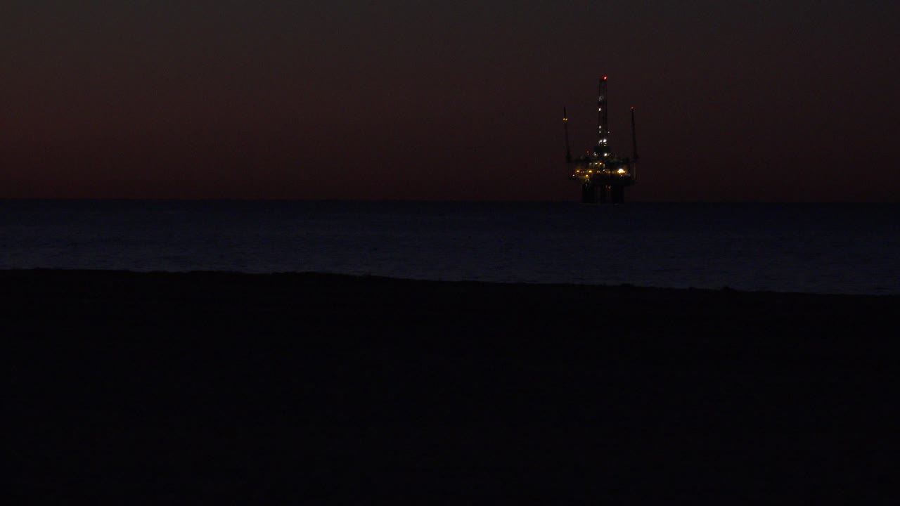 Huntington Beach twilight frame with a dark beach foreground, low ocean band, and a lit offshore platform or marine structure near the left side of the horizon, with no visible people.