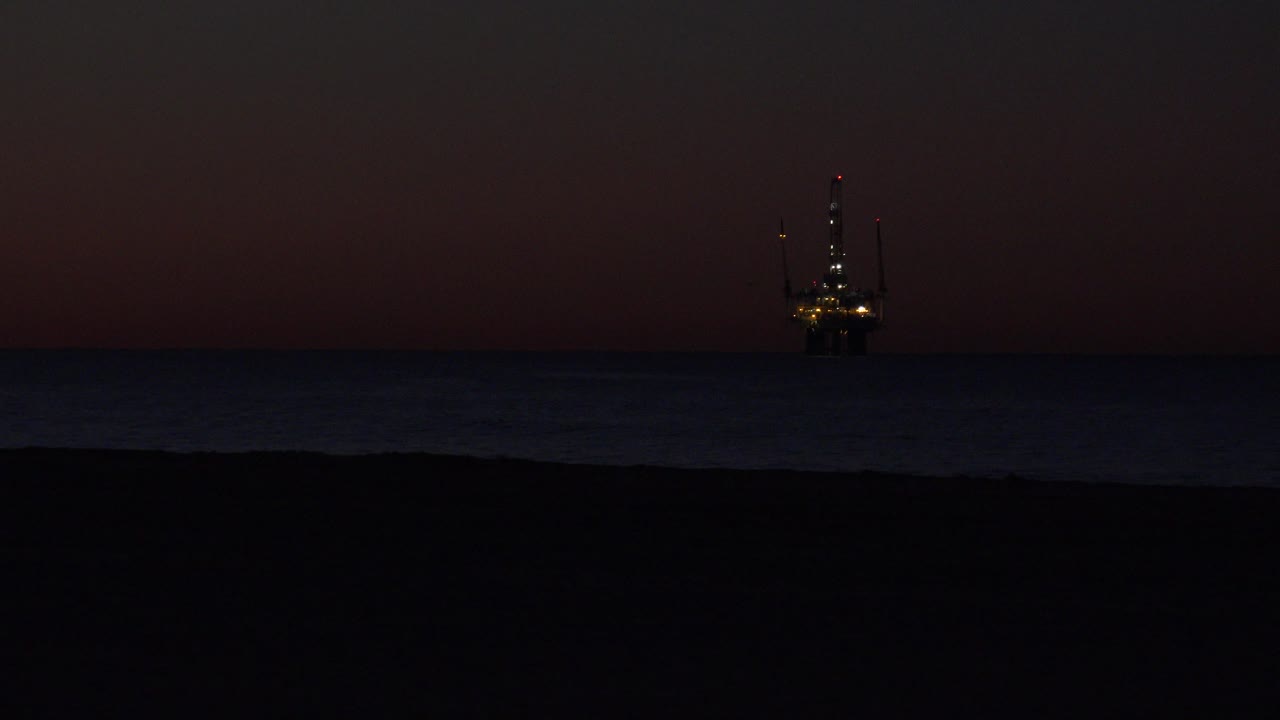 Huntington Beach at night with black sand in the foreground, dim ocean water, a small lit offshore structure near the center-left horizon, and no visible people.