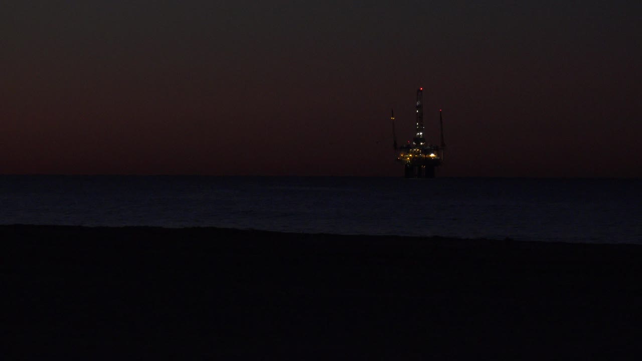Huntington Beach night shoreline with a broad dark foreground, faint waves, and the lit offshore structure closer to the center of the horizon, with no visible people.