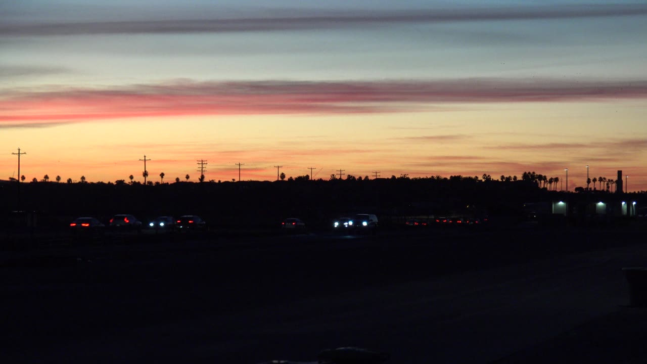 At 0:00, Huntington Beach sunset colors spread above a dark roadway with a few headlights, taillights, utility poles, distant palms, and no visible people.