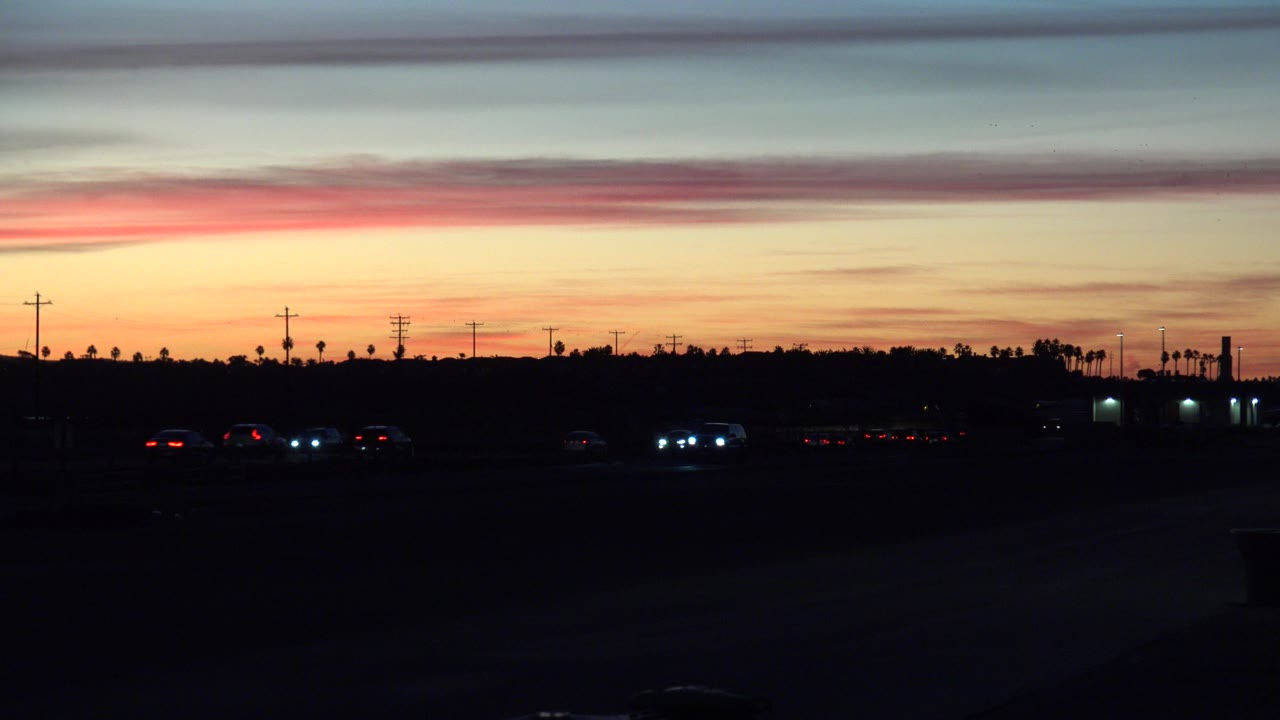 At 0:01, a few vehicles sit low in the dark foreground while orange and pink bands brighten the Huntington Beach horizon, with utility poles and no visible people.