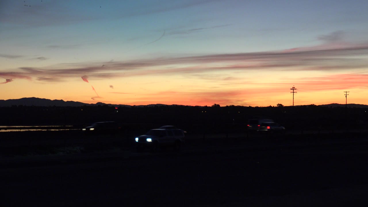 Opening frame of Huntington Beach traffic at sunrise, with car headlights in the dark foreground, utility poles near the horizon, distant hills, and no visible people.