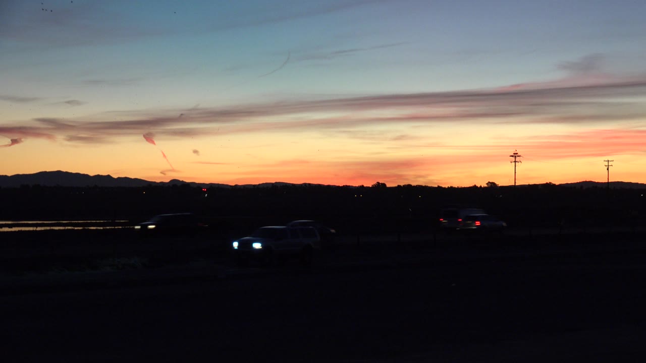 Huntington Beach sunrise frame with a vehicle's headlights lower in frame, another car silhouette farther right, dark roadside foreground, utility poles, distant hills, and no visible people.