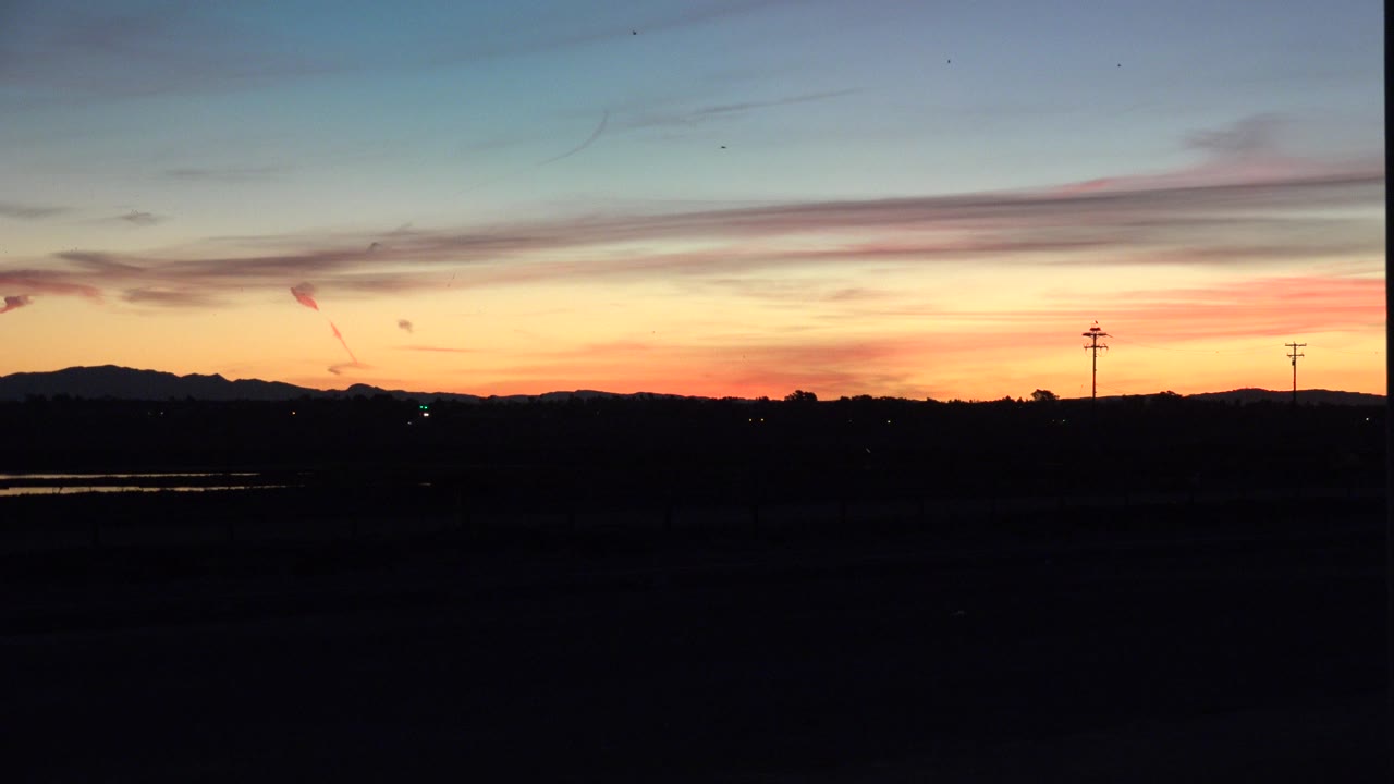 Second frame of the Huntington Beach sunset, with thin horizontal clouds stretched across the blue and orange sky above a nearly black foreground and no visible people.