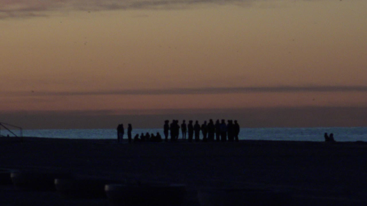 A distant group of silhouetted people stands and sits on Huntington Beach at sundown, with a dark beach foreground and a low ocean horizon behind them.