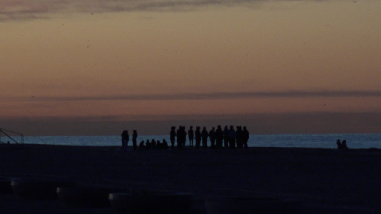 Silhouetted people remain spread along Huntington Beach, with seated figures to the left, a standing cluster to the right, and the ocean catching a narrow band of light.