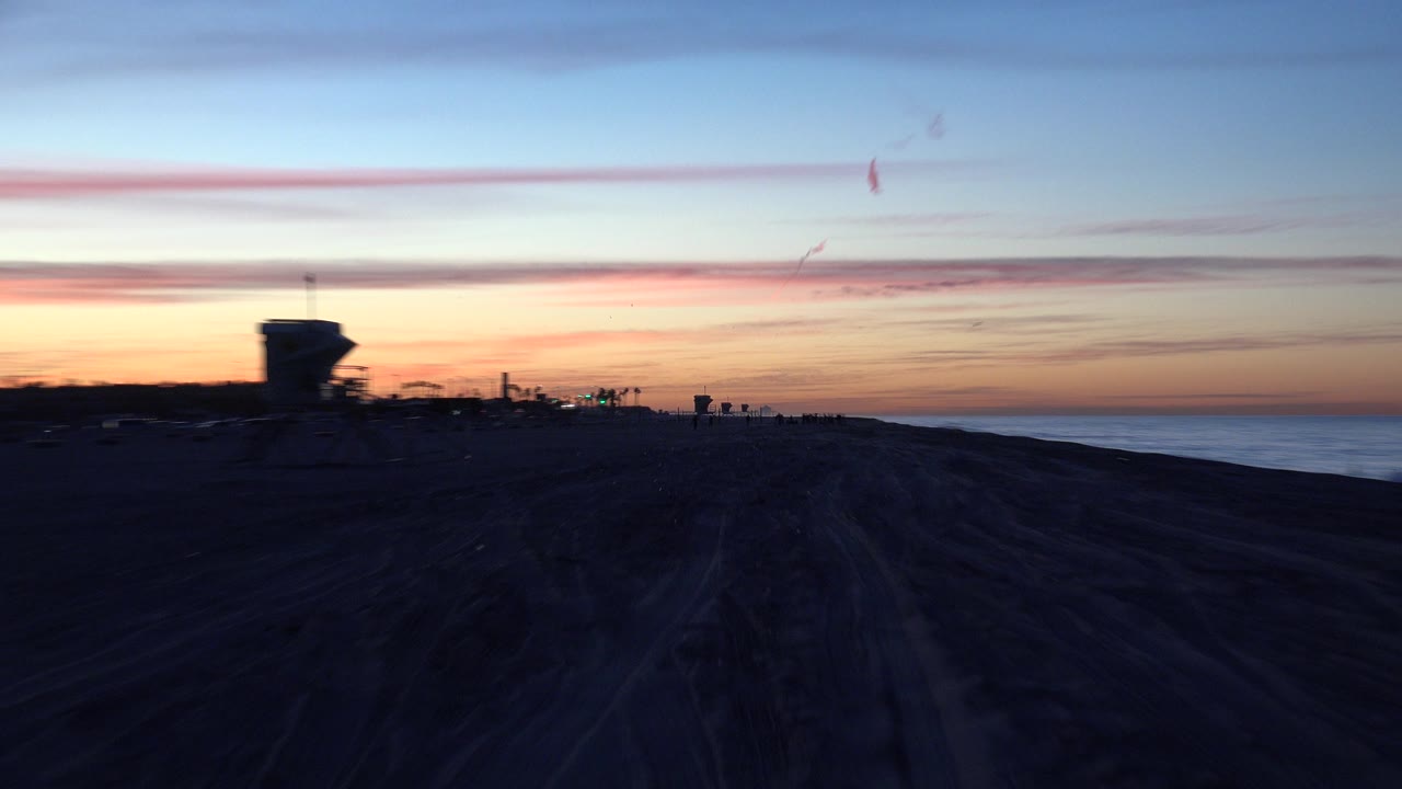 Huntington Beach at dusk with dark sandy foreground, a distant tower-shaped silhouette near the horizon, orange and blue sky, and no visible people.