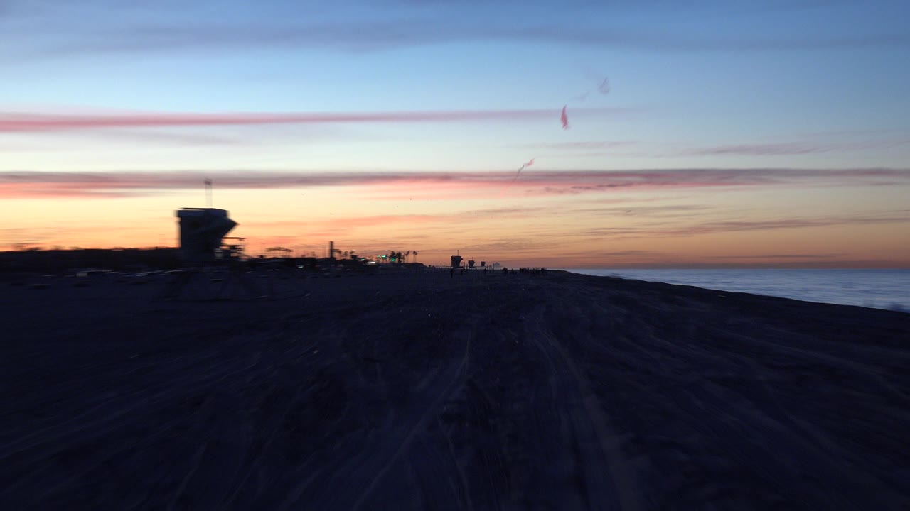 Huntington Beach sunset view with rutted sand in shadow, a thin ocean line to the right, warm light at the horizon, and no visible people.