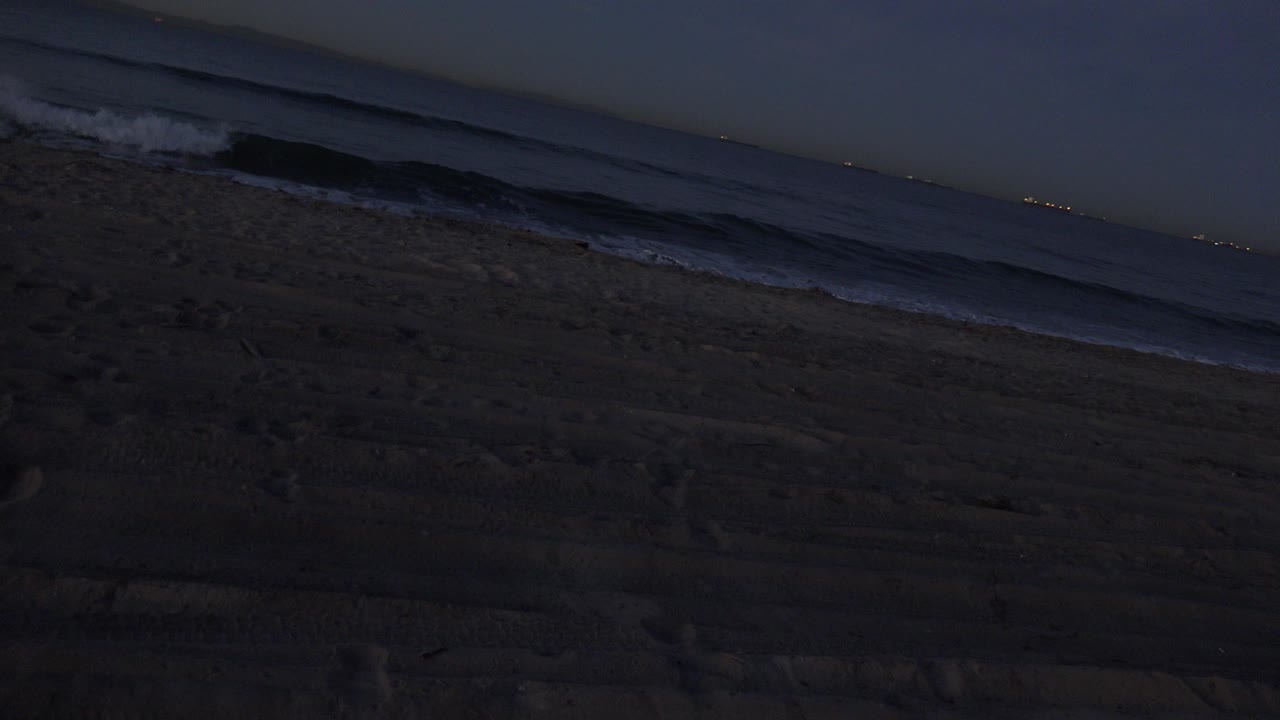 Opening frame of Huntington Beach at night with dark sand in the foreground, low surf bands in the distance, faint lights on the horizon, and no visible people.