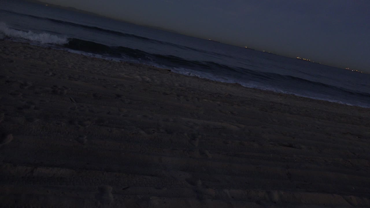 Huntington Beach night frame with a small white break forming on the left side of the surf line, dark sand below, distant lights, and no visible people.