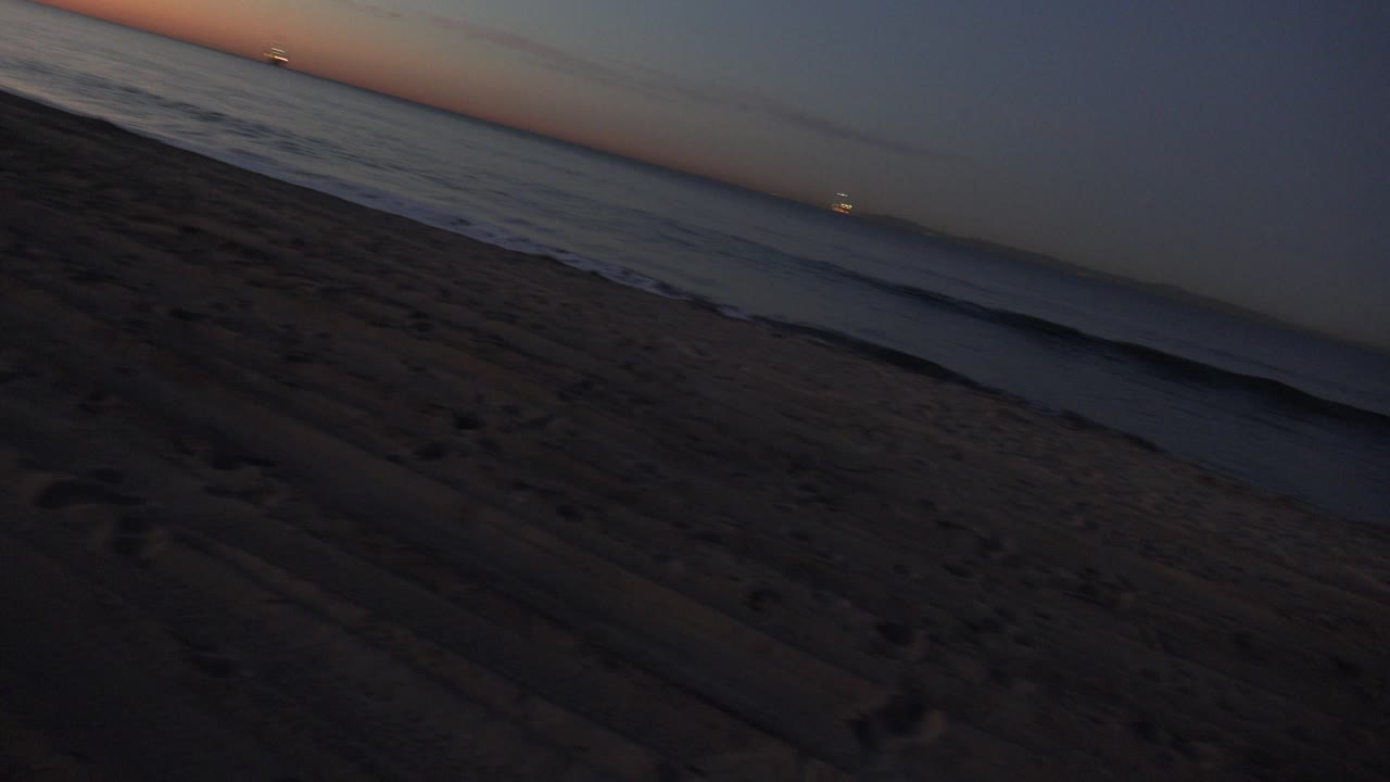 Tilted ground-level view of Huntington Beach sand and shoreline at sunset, with small waves on the left and no visible people.